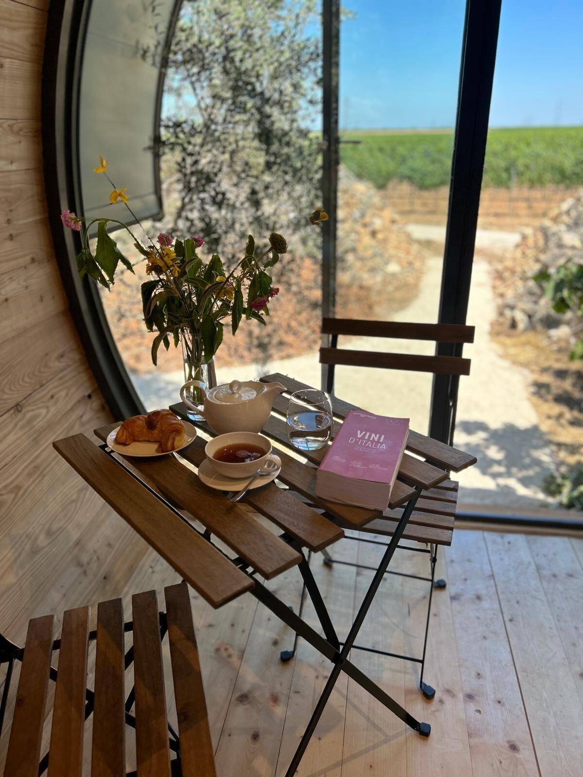 A wooden table with a book, a bowl of soup, a cup of tea, and a croissant.