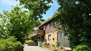 A rustic wooden house with a balcony, surrounded by lush greenery and a gravel driveway. - Les Vignes (Durbuy)