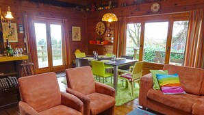 A cozy room with wooden walls, a dining table with chairs, a sofa with colorful cushions, and a large window with curtains. - Les Vignes (Durbuy)