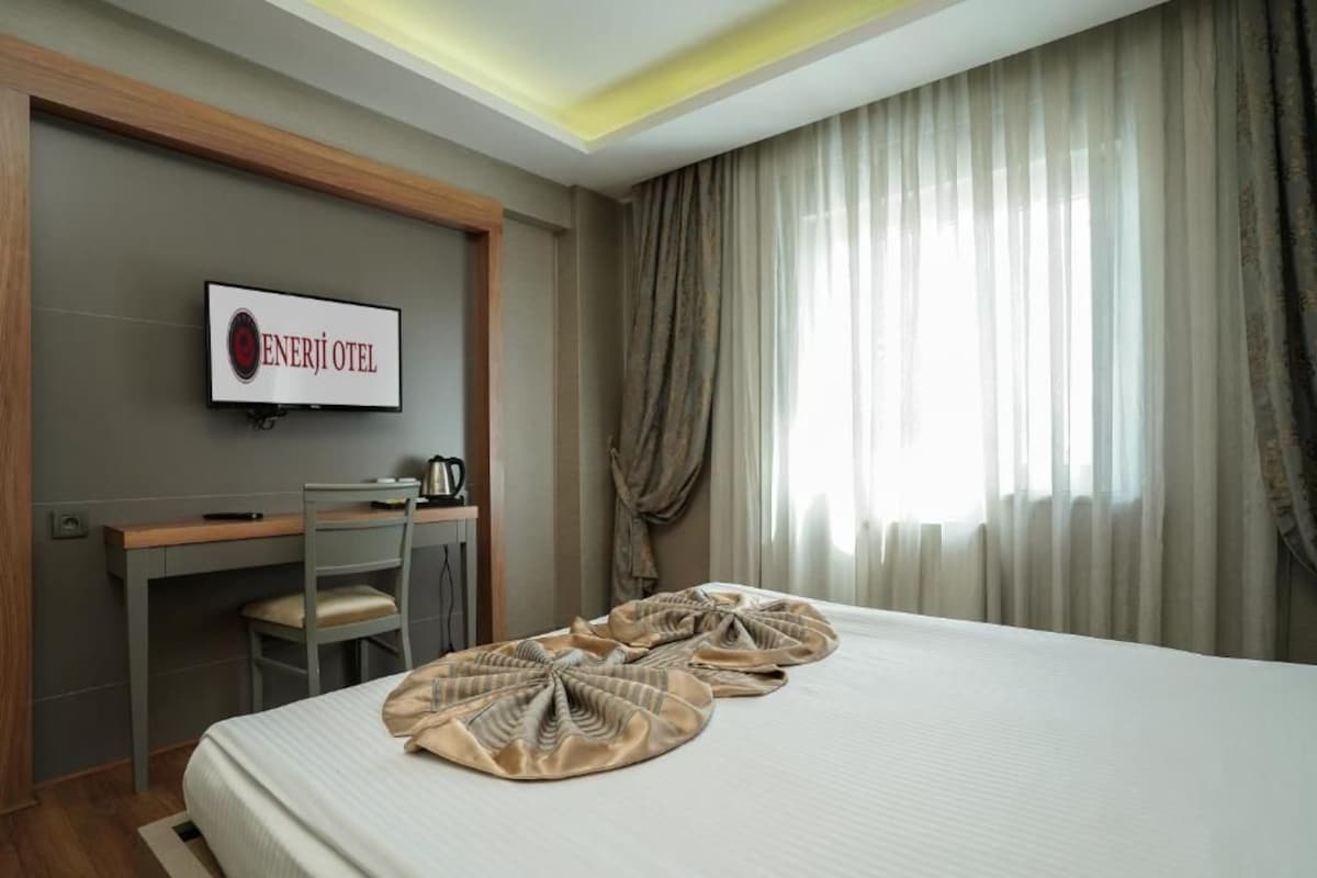 A hotel room with a bed, a desk, a chair, a TV displaying 'ENERJI OTEL', and a window with curtains.
