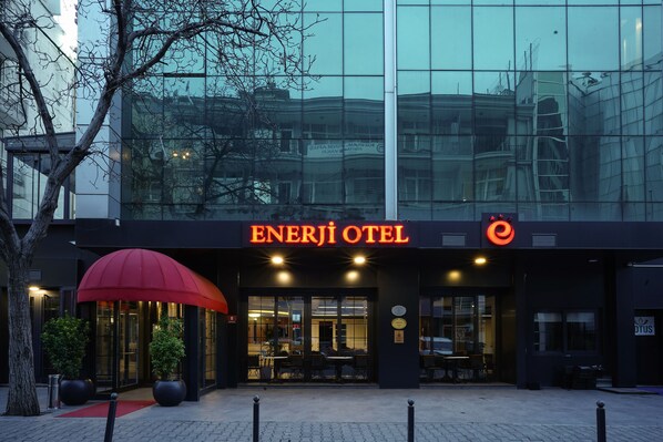 The exterior of Enerji Hotel with a red umbrella, glass facade, and outdoor seating area.