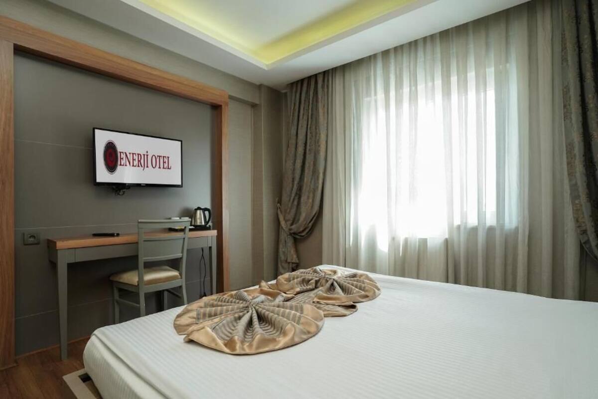 A hotel room with a bed, a desk, a chair, a TV displaying 'ENERJI OTEL', and a window with curtains.