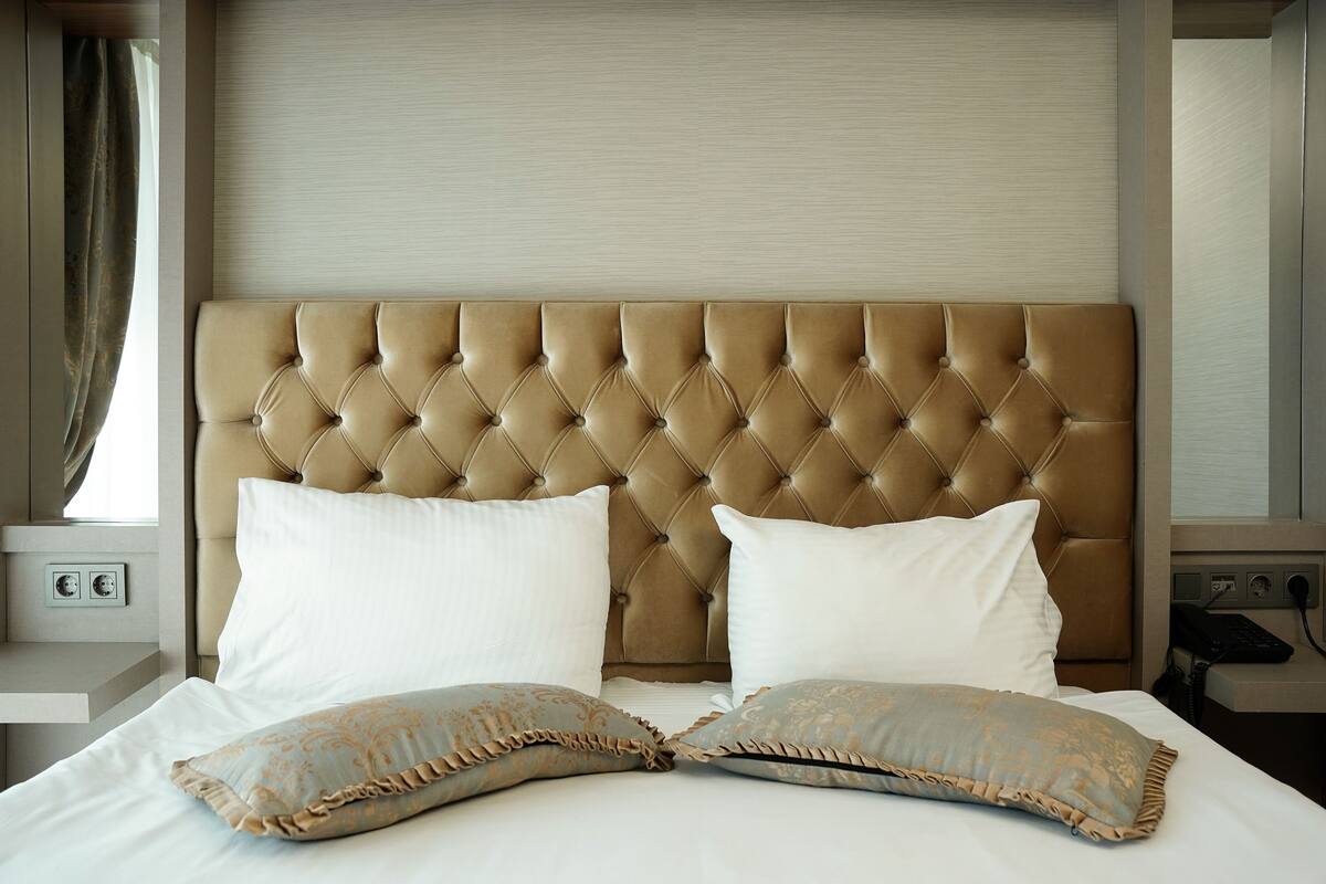 A hotel room with a tufted headboard, two white pillows, and decorative pillows with a ruffled edge.