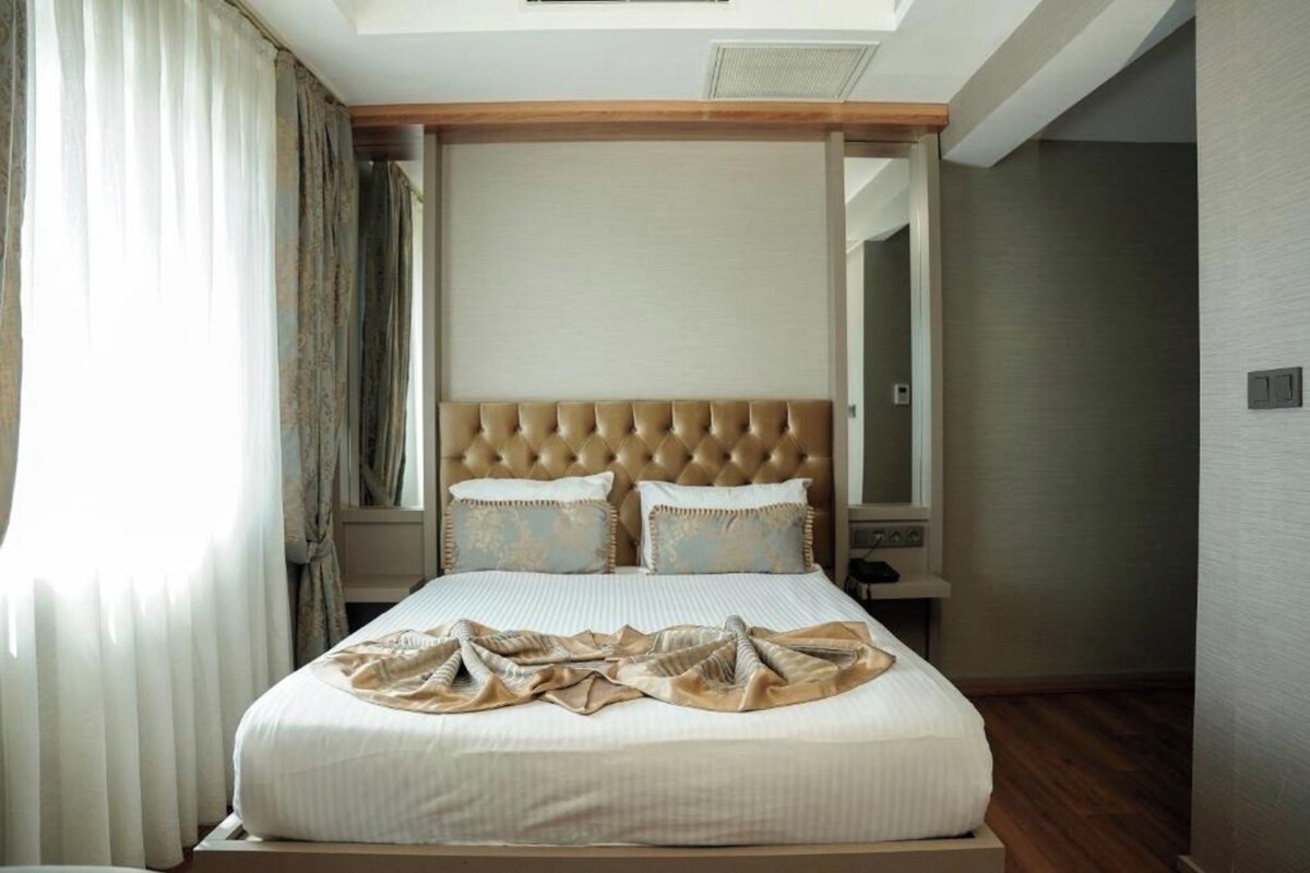 A bedroom with a tufted headboard, a bed with a white bedspread, two pillows, a bedside table, and a window with curtains.