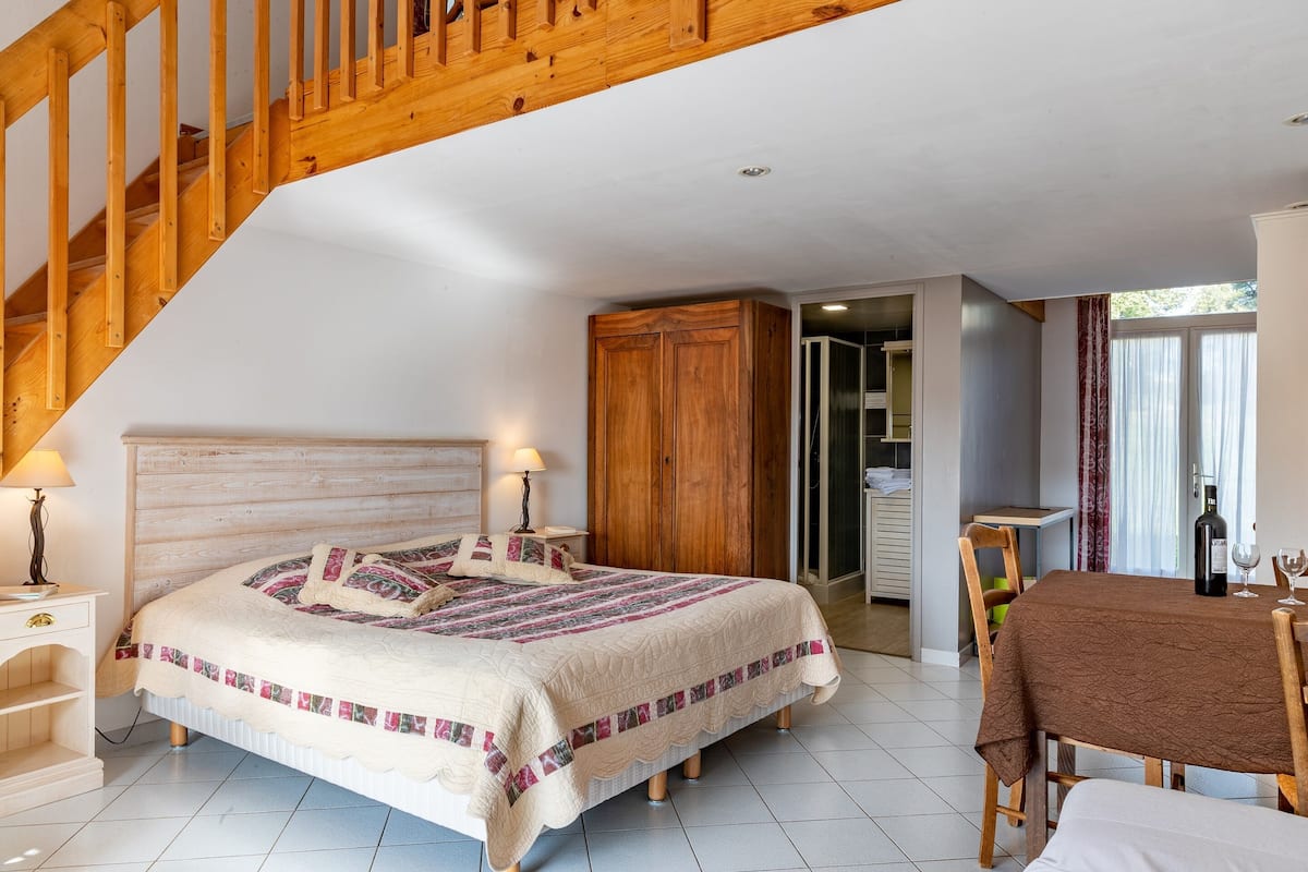 A bedroom with a wooden staircase, a bed with a quilt, a wooden wardrobe, a dining table with a tablecloth, and a bottle of wine.