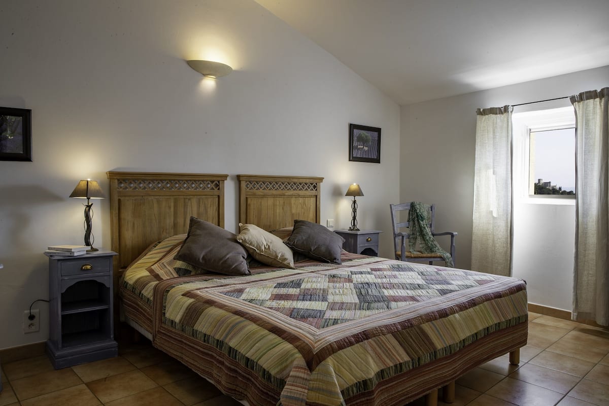 A bedroom with a large bed, two bedside tables, a nightstand, a lamp, a framed picture, and a window with curtains.