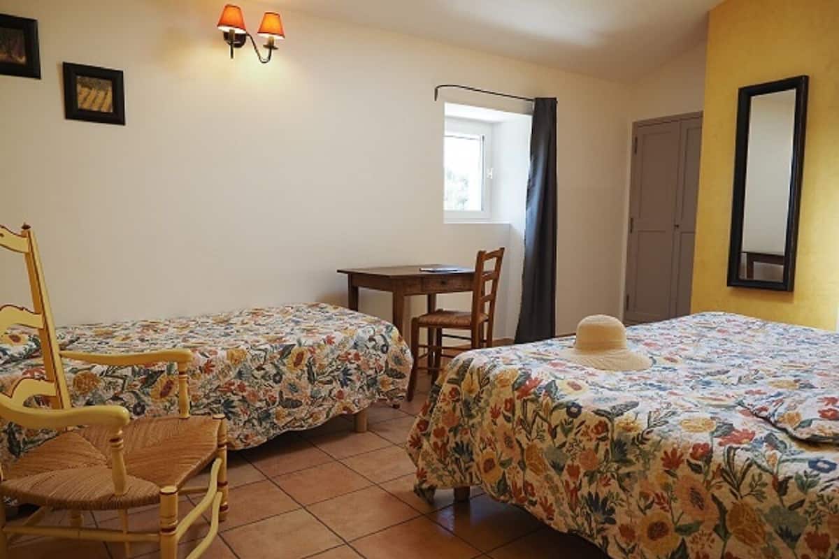 A room with two single beds, a wooden chair, a small table, a window with curtains, and a wall-mounted lamp.