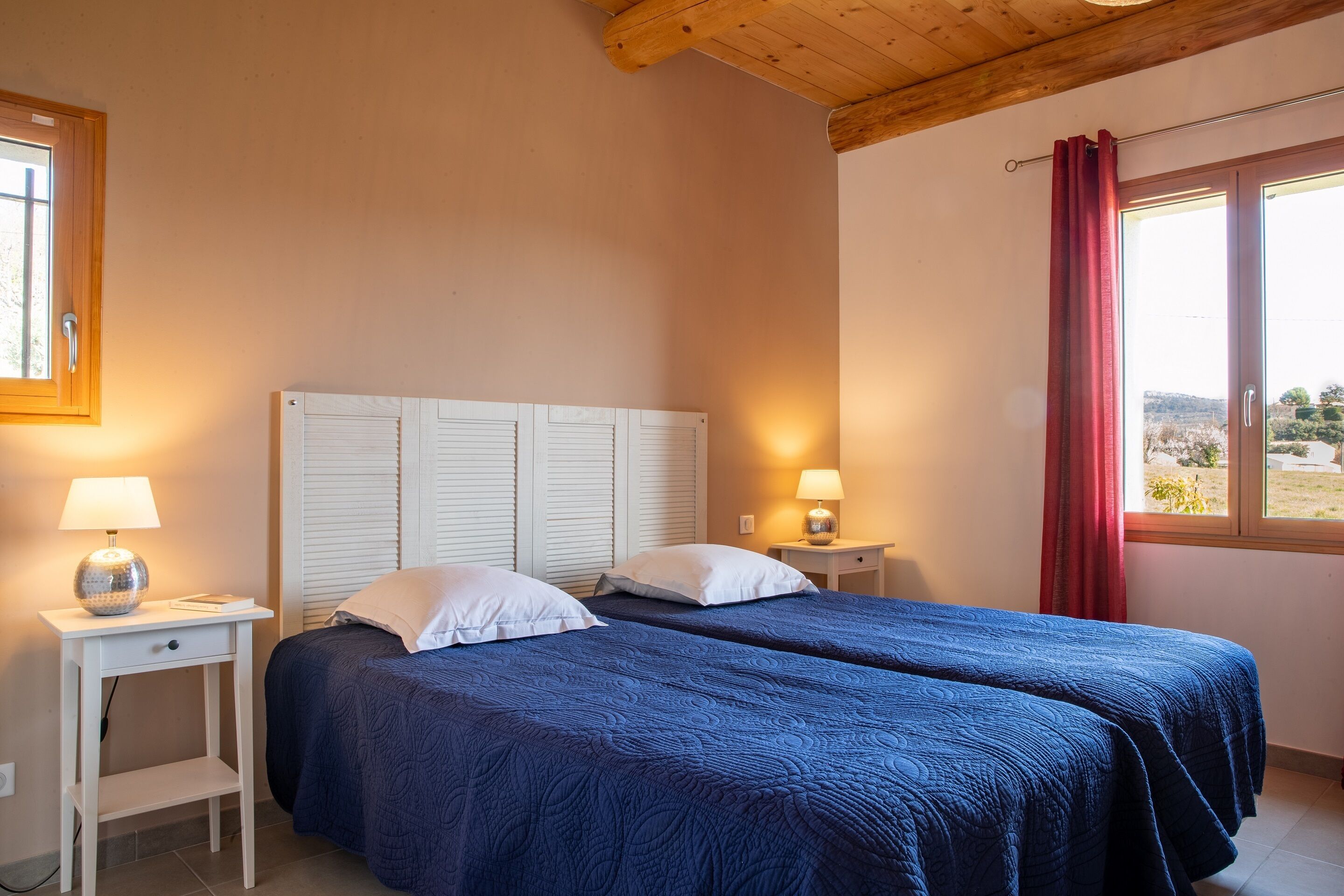 Een slaapkamer met twee bedden, witte hoofdeinden, nachtkastjes met lampen en een raam met een rood gordijn.