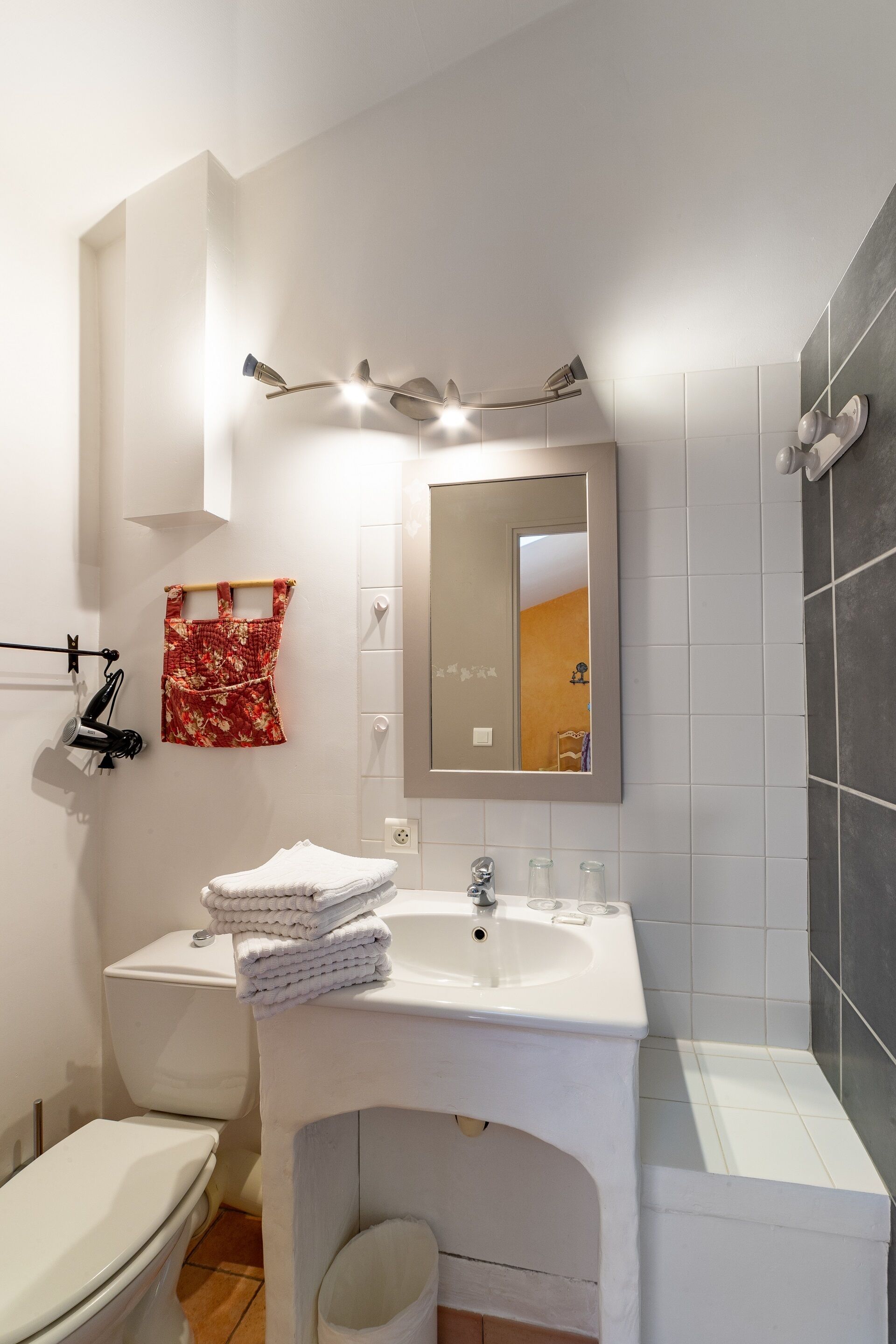 Casa de banho com lavatório branco, espelho e sanita. Tem um porta-toalhas com toalhas dobradas e uma toalha vermelha com padrão floral na parede.