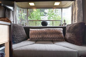 A cozy seating area with a cushioned bench and two large pillows, a small round table with a Buddha statue, and a window with a view of greenery and a building outside. - Seattle Glamping - Zen Den (Seattle)