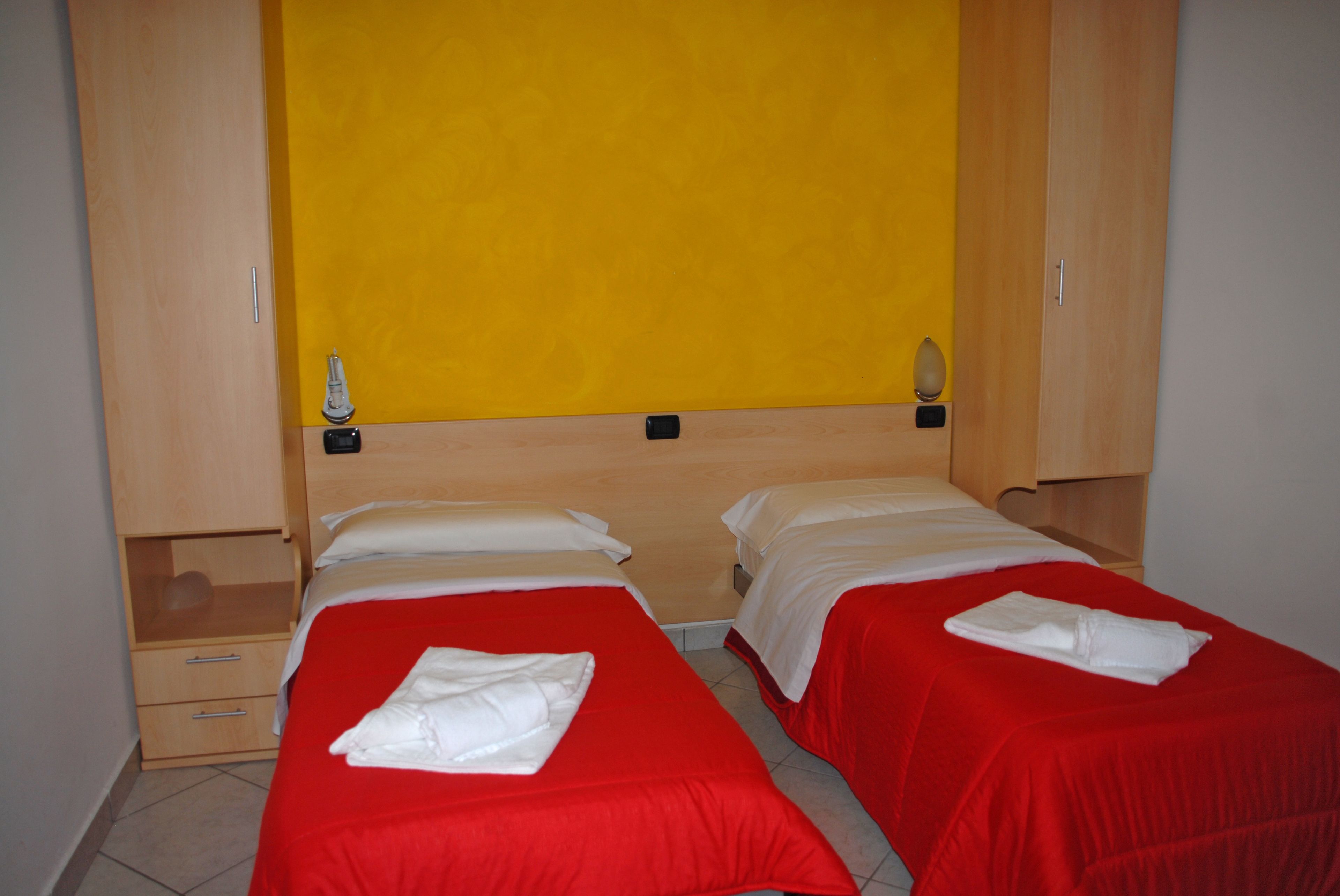 A hotel room with two single beds, each with a red bedspread and white pillows, and a wooden wardrobe with a yellow accent wall.