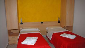 A hotel room with two single beds, each with a red bedspread and white pillows, and a wooden wardrobe with a yellow accent wall.