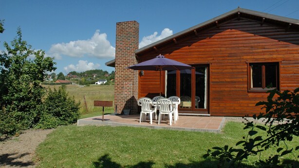 Una cabaña de madera con chimenea de ladrillo, un patio con sillas blancas y vistas del paisaje circundante.