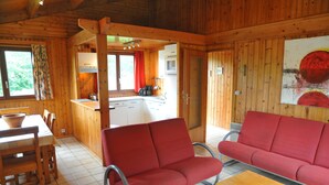 Interior de una cabaña de madera con comedor, sofá rojo y cocina visible a través de una puerta abierta.
