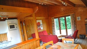 Interior de una cabaña de madera con cocina, sillas rojas, una mesa con un bol y ventanas amplias.