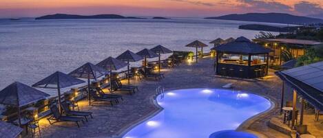 A pool area with lounge chairs and umbrellas, surrounded by a stone deck, overlooking a body of water at sunset.