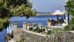 A stone-walled terrace with outdoor seating overlooking a body of water, surrounded by greenery and flags. - Blue Bay Resort Village (Andros)