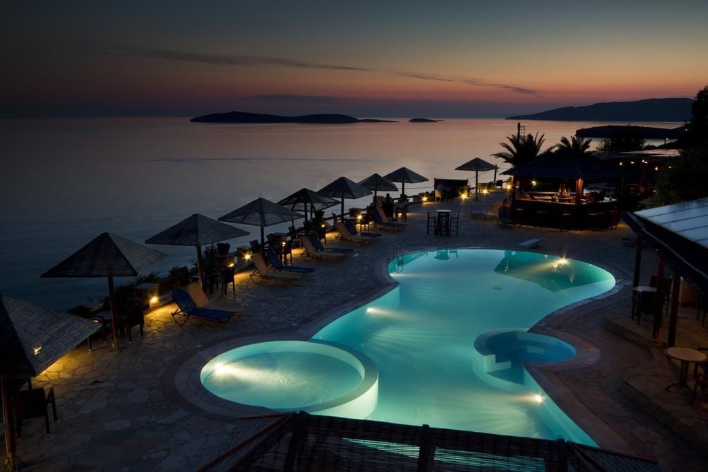 A pool area with lounge chairs and umbrellas, surrounded by a stone deck, overlooking a body of water at dusk.