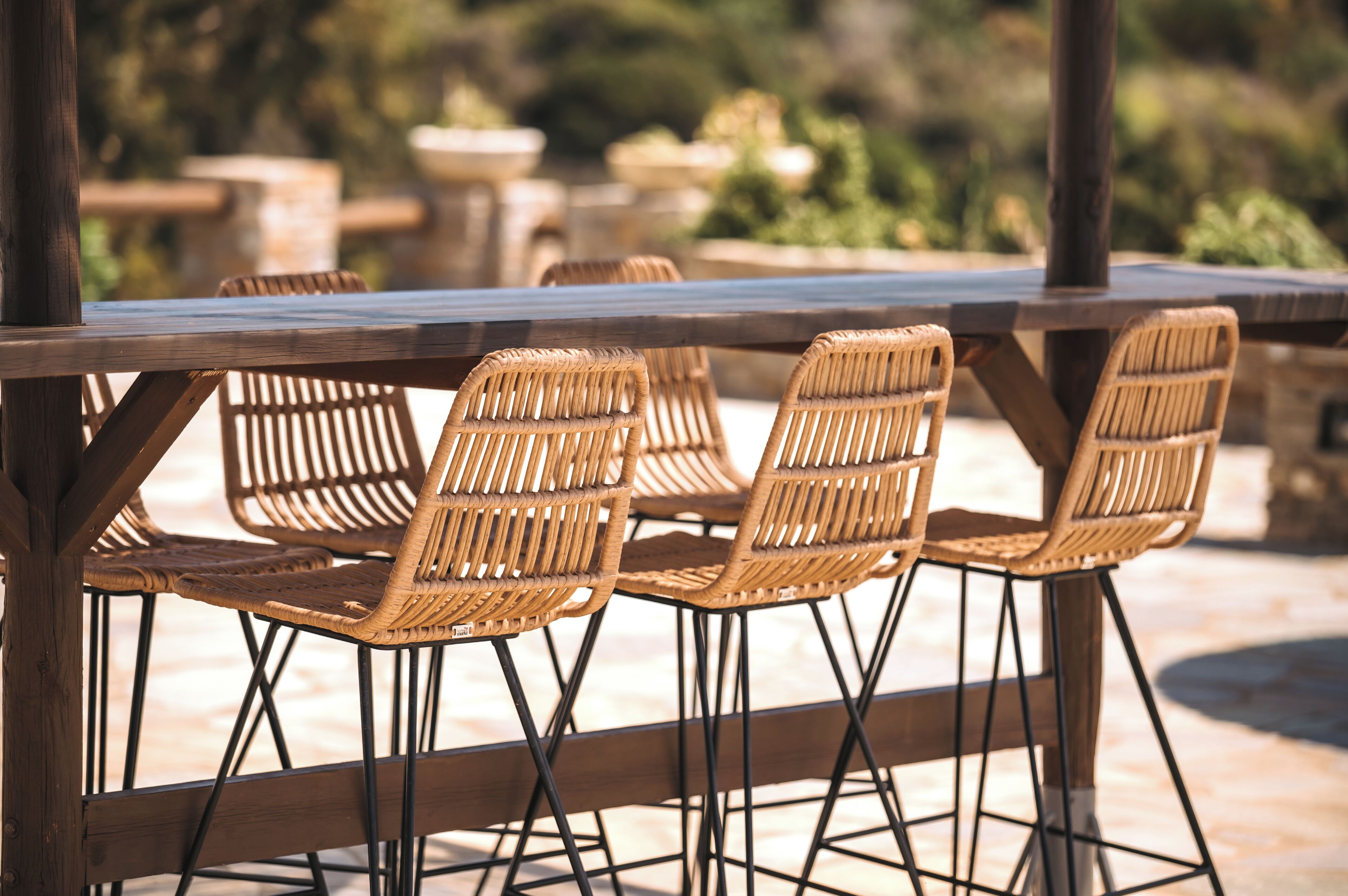 A wooden table with wicker chairs, set outdoors with a blurred background of greenery.