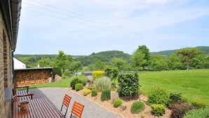 A patio with a wooden table and chairs, a brick wall, and a view of a green field and trees. - Wilgenhof (Hotton)