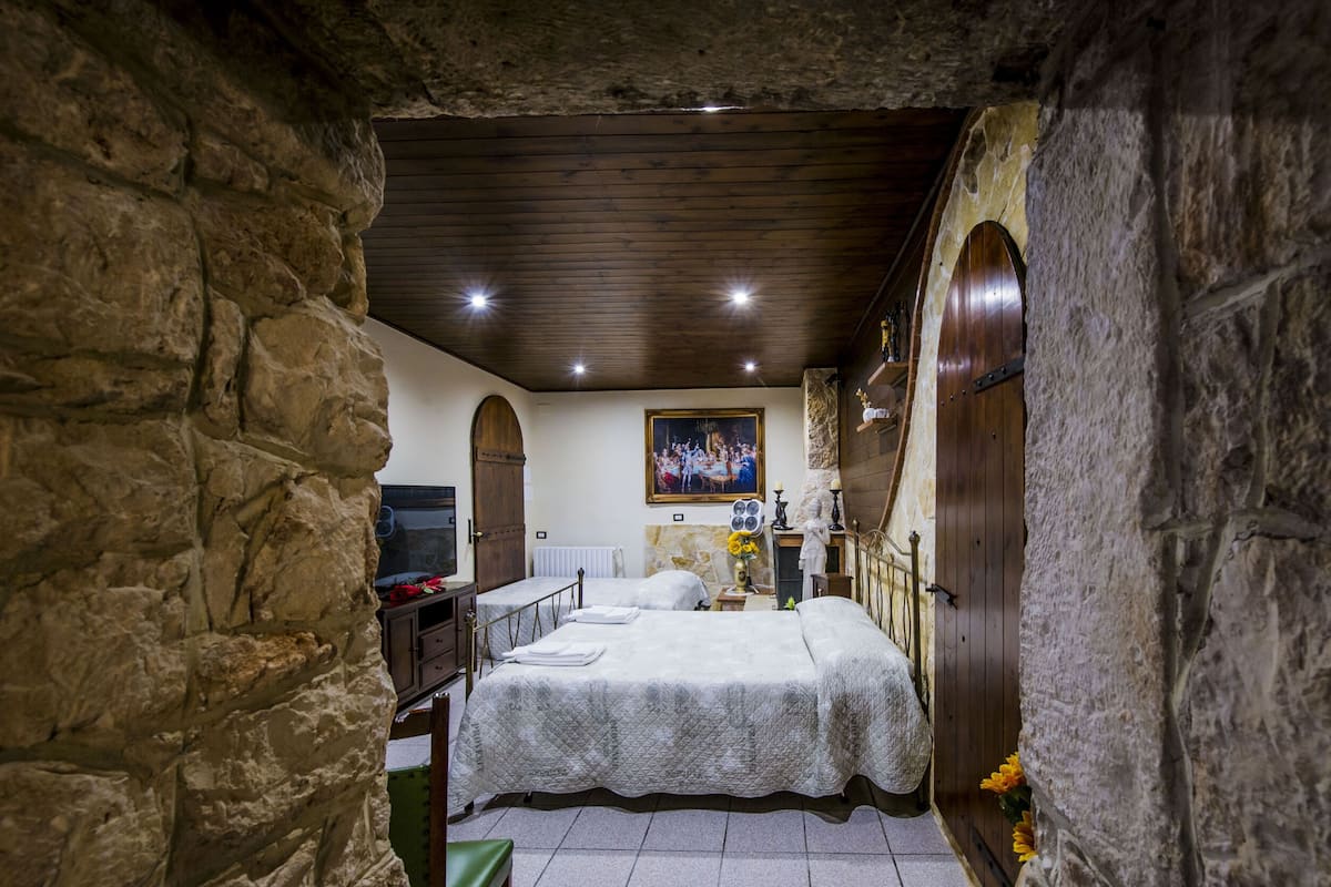 Une chambre à coucher rustique avec des murs en pierre, un plafond en bois, un lit recouvert d’une courtepointe, une commode, un miroir et un tableau accroché au mur.