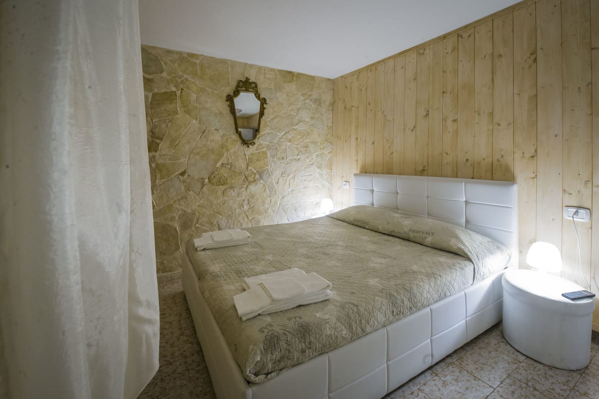 Une chambre avec un mur en pierre, un lambris en bois, un lit blanc et une table de chevet avec une lampe.