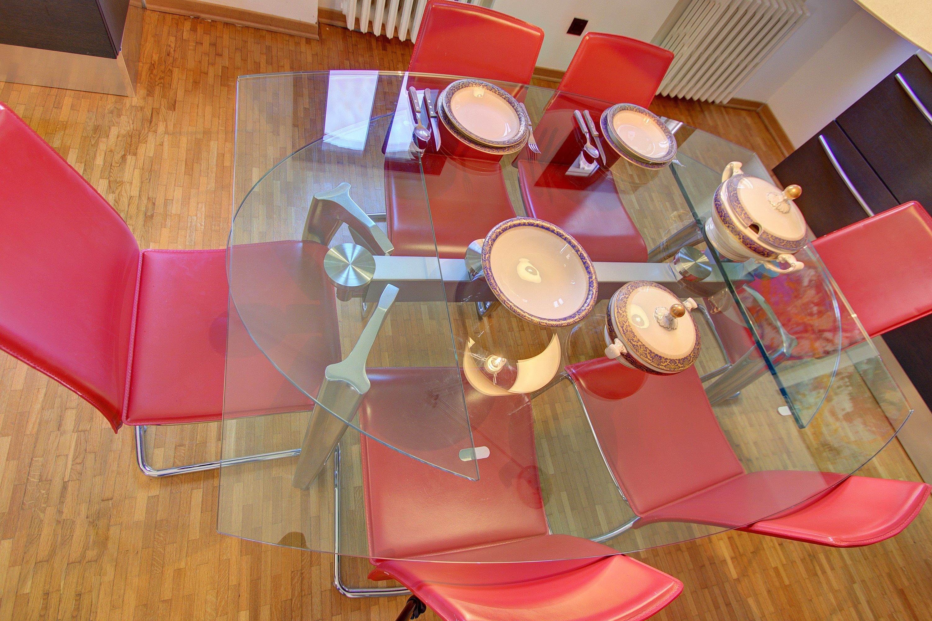 A round glass table with six red chairs, surrounded by a wooden floor.