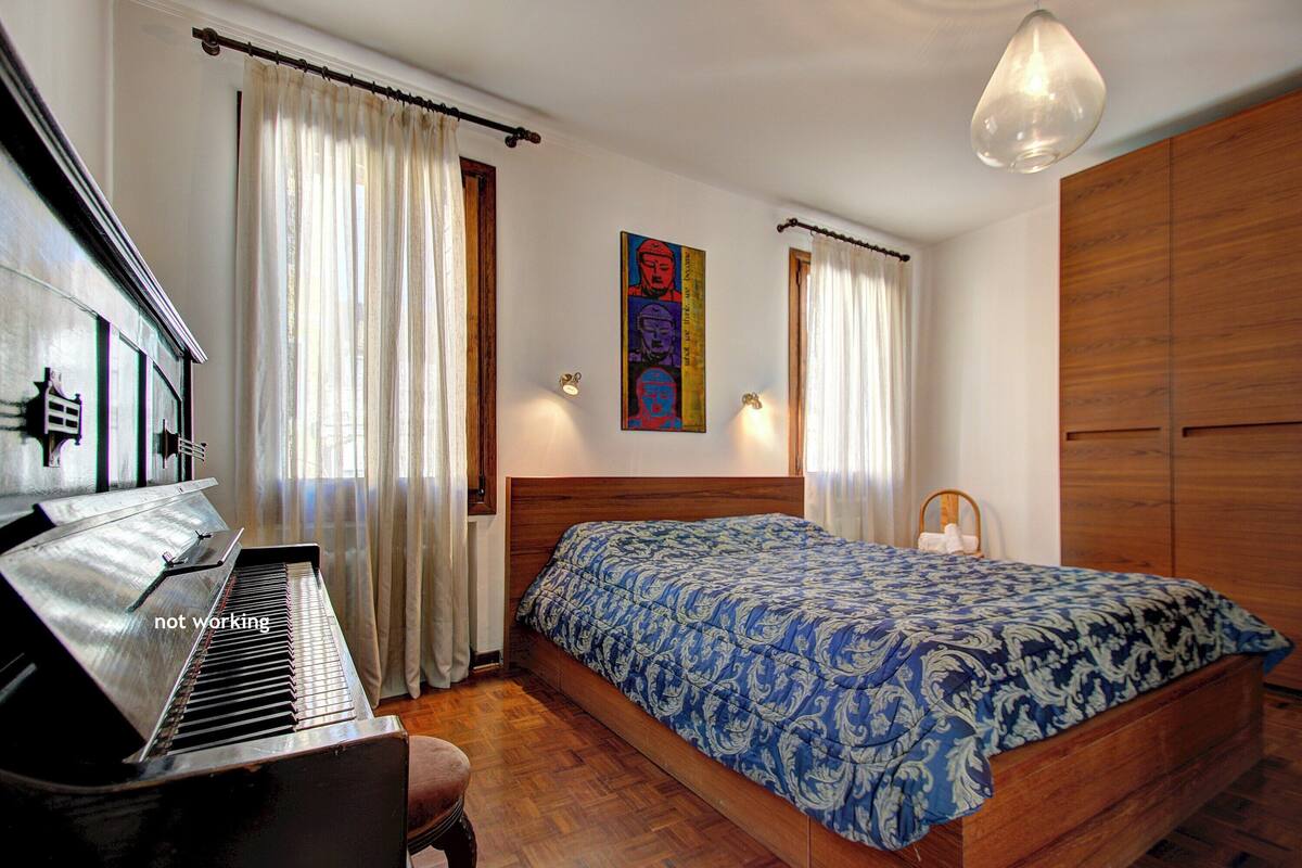 A bedroom with a piano, a bed with a patterned bedspread, and a wooden wardrobe.