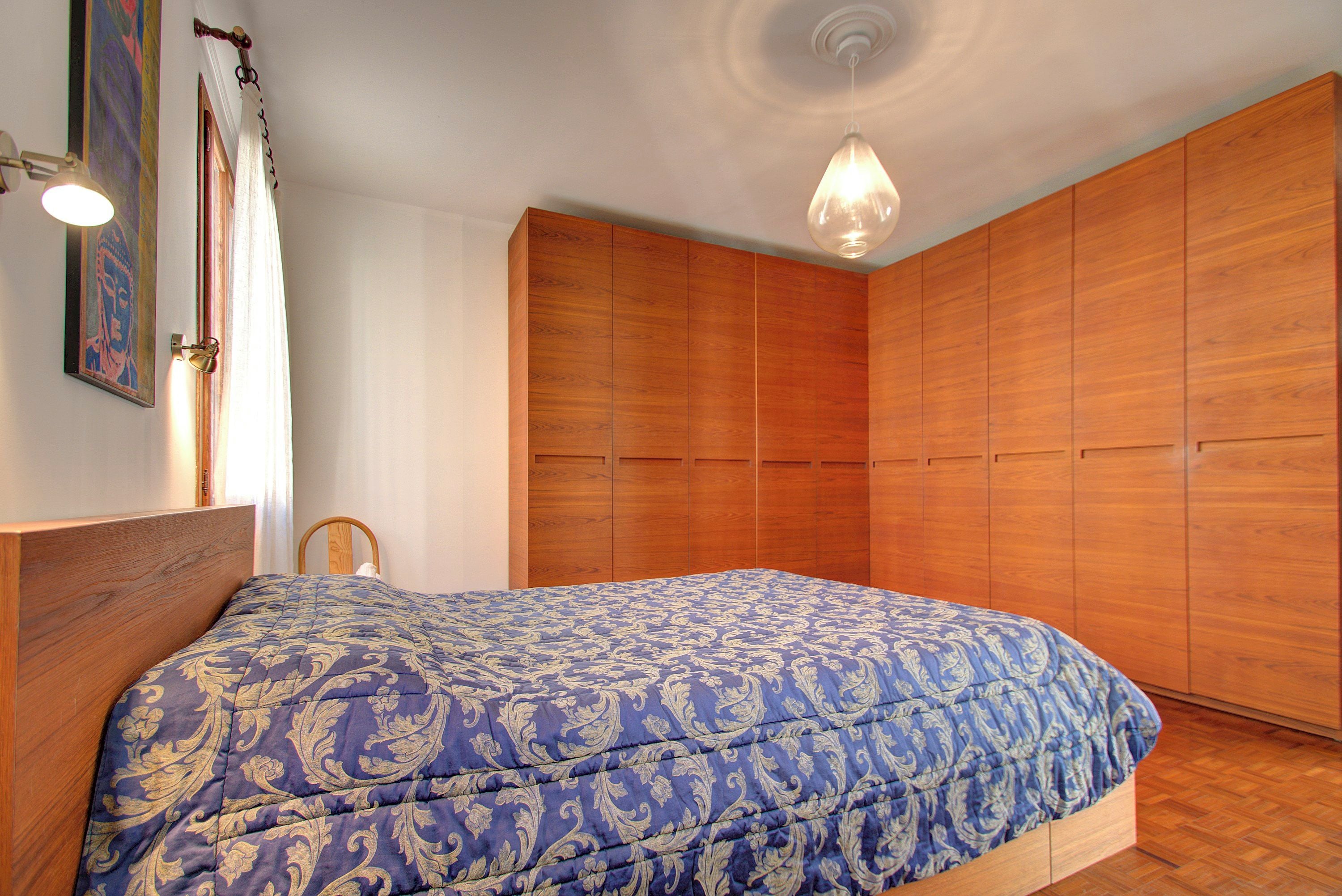 A bedroom with a wooden bed frame, a blue patterned bedspread, and a wooden wardrobe.
