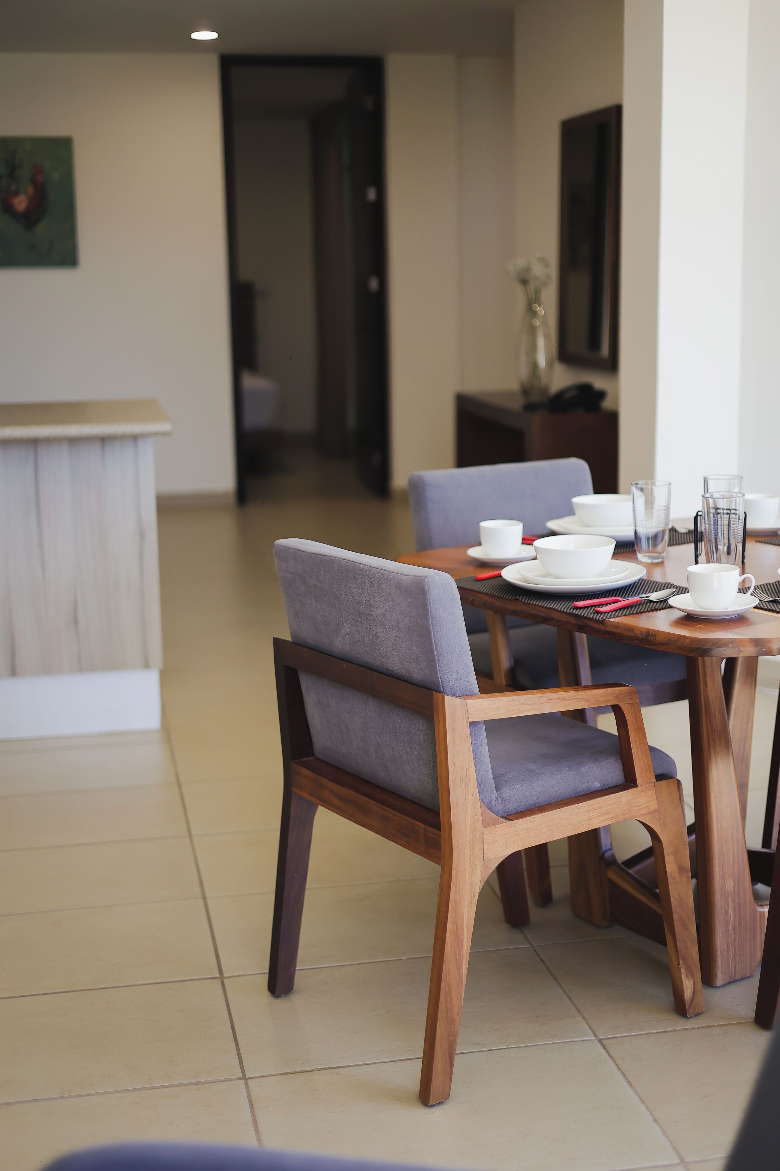 Comedor con una mesa de madera dispuesta para cuatro personas, incluyendo platos, tazones, vasos y cubiertos. Hay dos sillas con tapicería gris. El piso es de baldosas y la pared de color claro presenta un cuadro enmarcado.