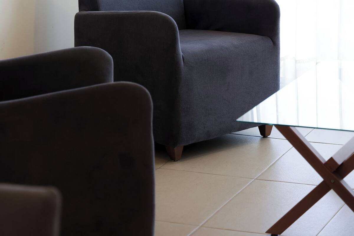 A modern living room with a dark grey armchair, a glass coffee table, and a view of a window with curtains.