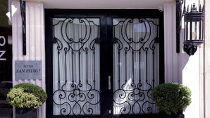 Ein Hotel Eingang mit einem Schild mit der Aufschrift "Suites San Pedro", zwei Blumentöpfen und verzierten schwarzen Metalltoren.