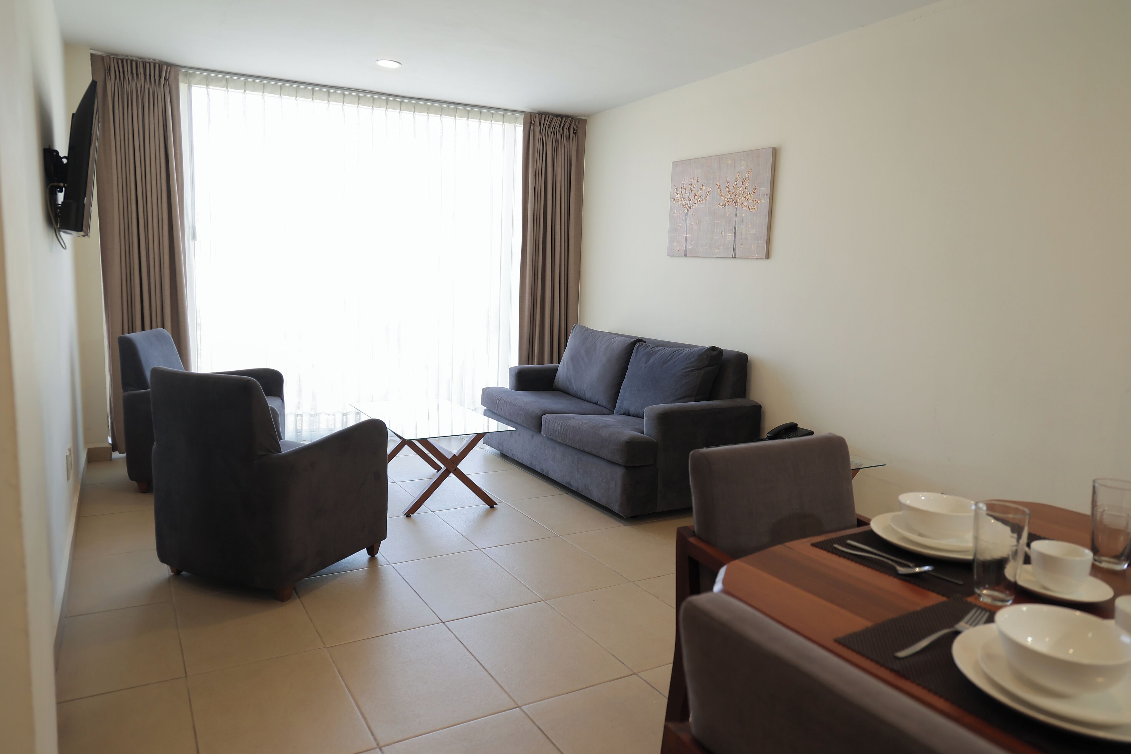 A modern living room with a grey sofa, a dining table set with plates and glasses, and a large window with curtains.