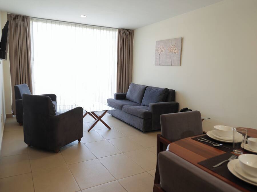 A modern living room with a grey sofa, a dining table set with plates and glasses, and a large window with curtains.