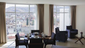 Ein Essbereich mit einem Tisch für vier Personen, großen Fenstern mit Vorhängen und Blick auf eine Stadtlandschaft mit Gebäuden und Bergen.