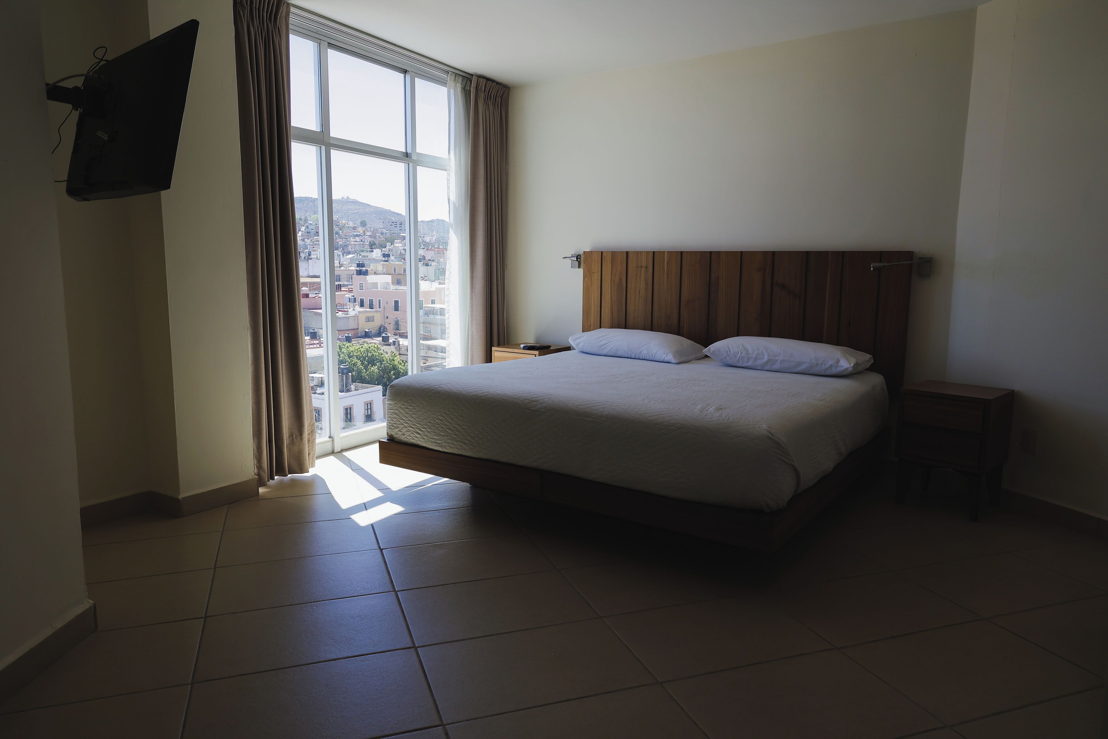 A hotel room with a large bed, a nightstand, a TV mounted on the wall, and a window with curtains.