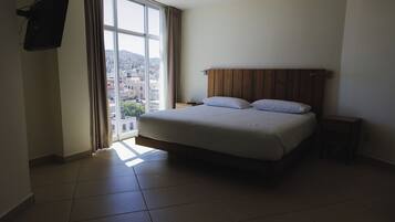 A hotel room with a large bed, a nightstand, a TV mounted on the wall, and a window with curtains.