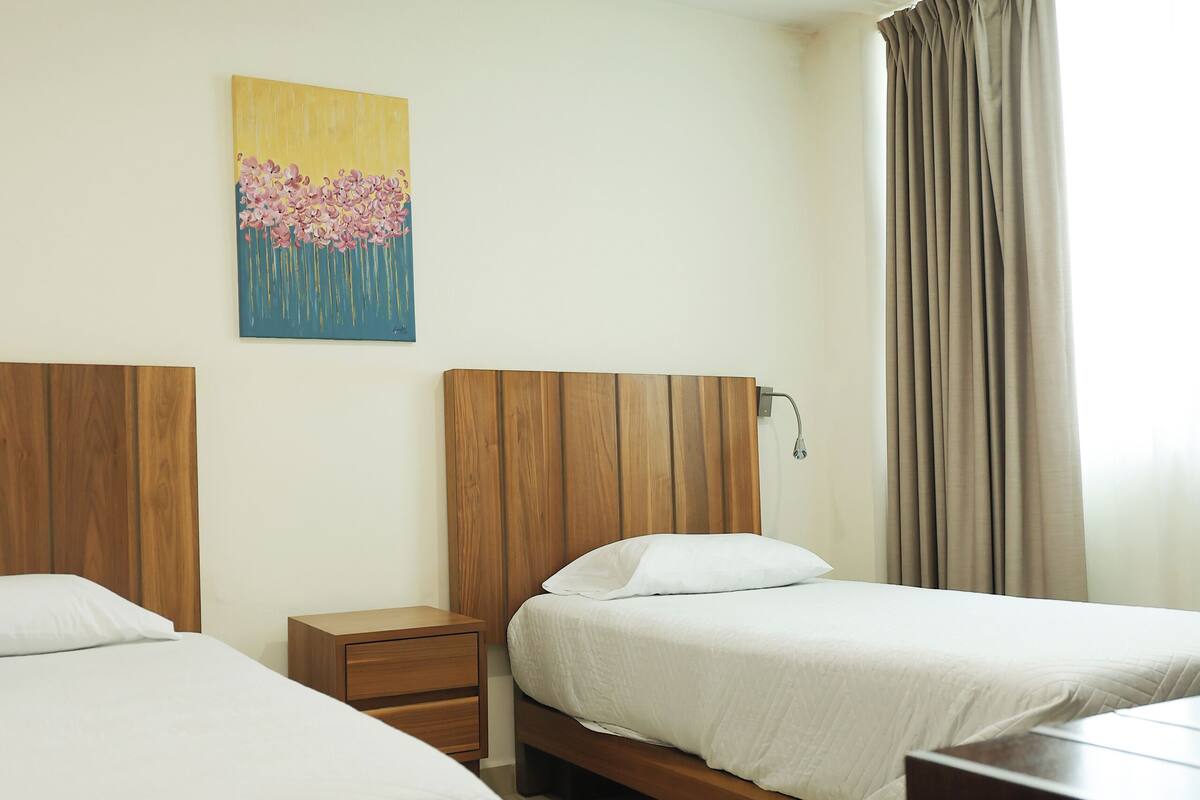 A hotel room with two beds, a nightstand, a painting on the wall, and a window with curtains.