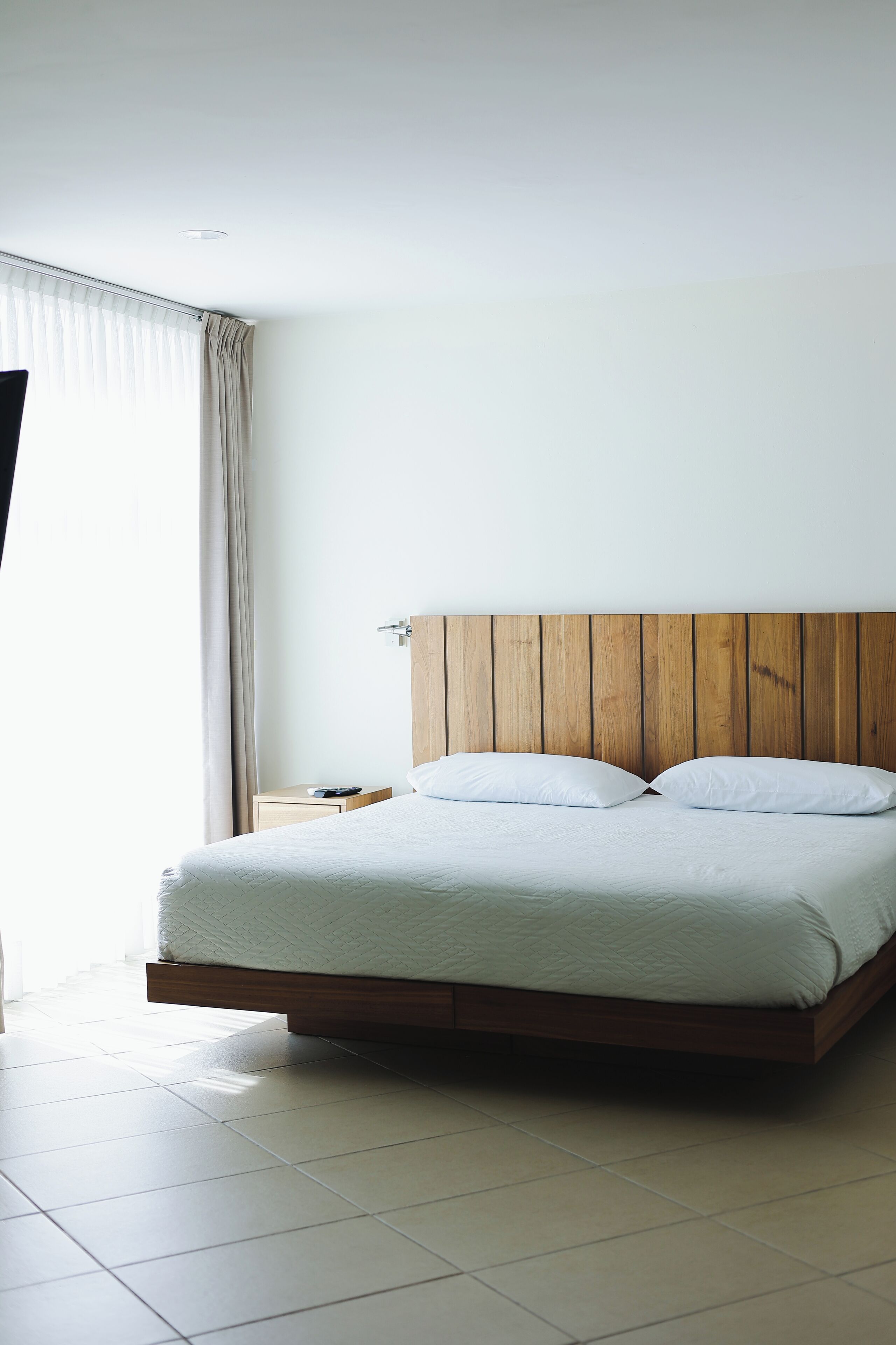 Una habitación de hotel moderna con cabecera de madera, cama con ropa blanca, mesita de noche y ventana con cortinas.