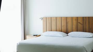Una habitación de hotel moderna con cabecera de madera, cama con ropa blanca, mesita de noche y ventana con cortinas.