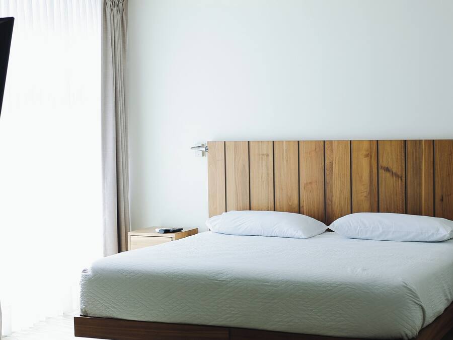 Una habitación de hotel moderna con cabecera de madera, cama con ropa blanca, mesita de noche y ventana con cortinas.
