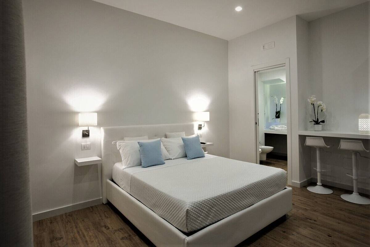 A modern bedroom with a large bed, bedside lamps, a small bedside table, and a bathroom visible through an open door.