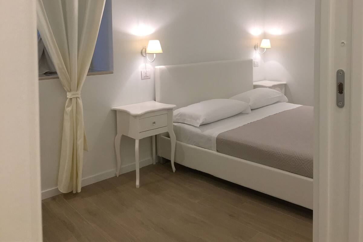 A bedroom with a bed, two bedside tables, a nightstand, a curtain, and wall-mounted lights.