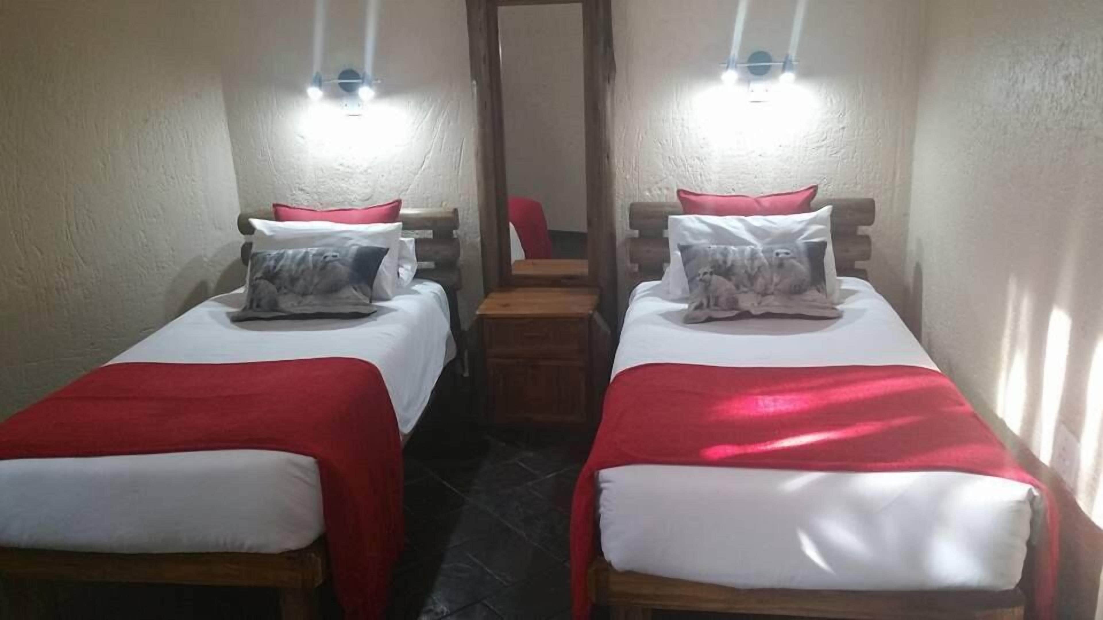 Una habitación con dos camas individuales, cada una con un edredón rojo y una almohada. Hay mesitas de noche con lámparas y un aparador de madera en el centro.