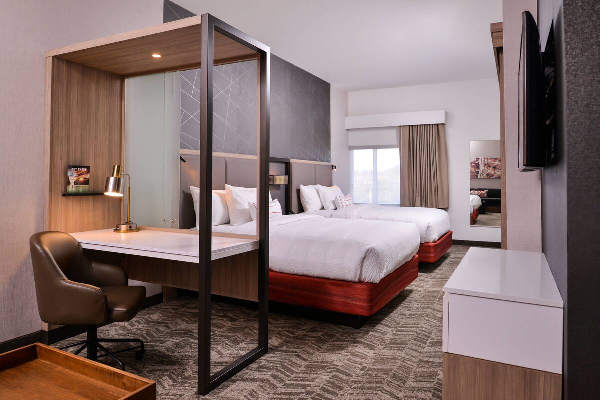 A hotel room with a desk, two beds, a TV, and a mirror.