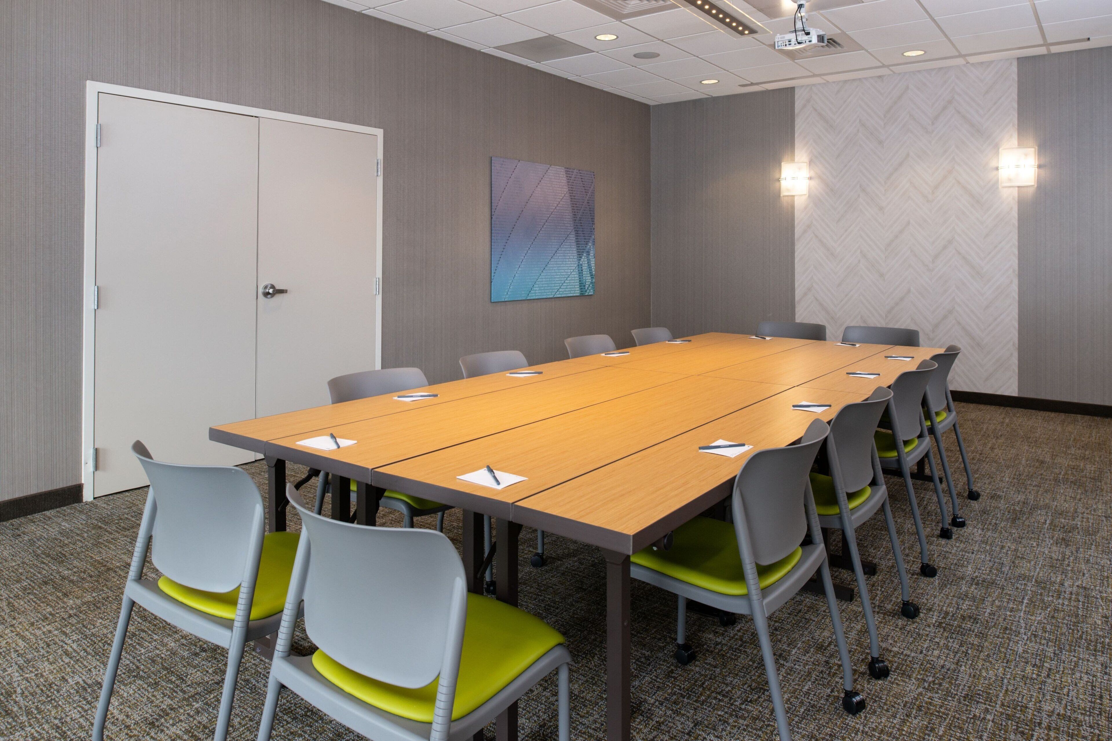 A conference room with a long table, grey chairs with green cushions, a white door, and a patterned wall.