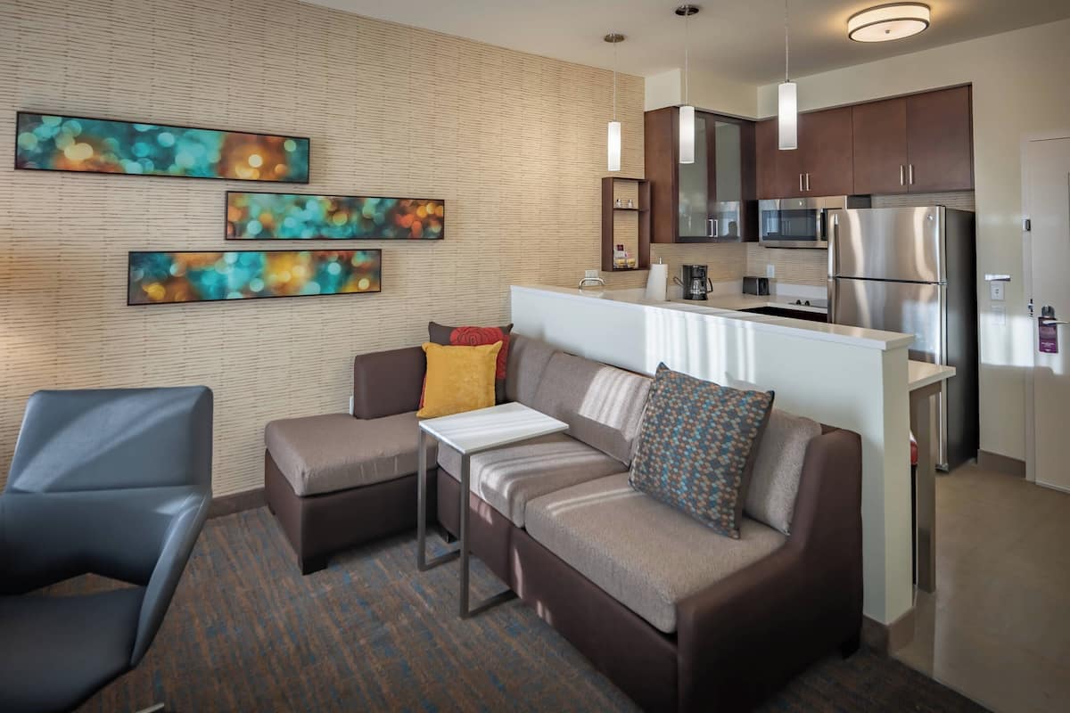 A modern living room with a sofa, ottoman, and a small table. There is a kitchen area with a refrigerator, microwave, and cabinets. The room has a wooden wall and a hanging light fixture.