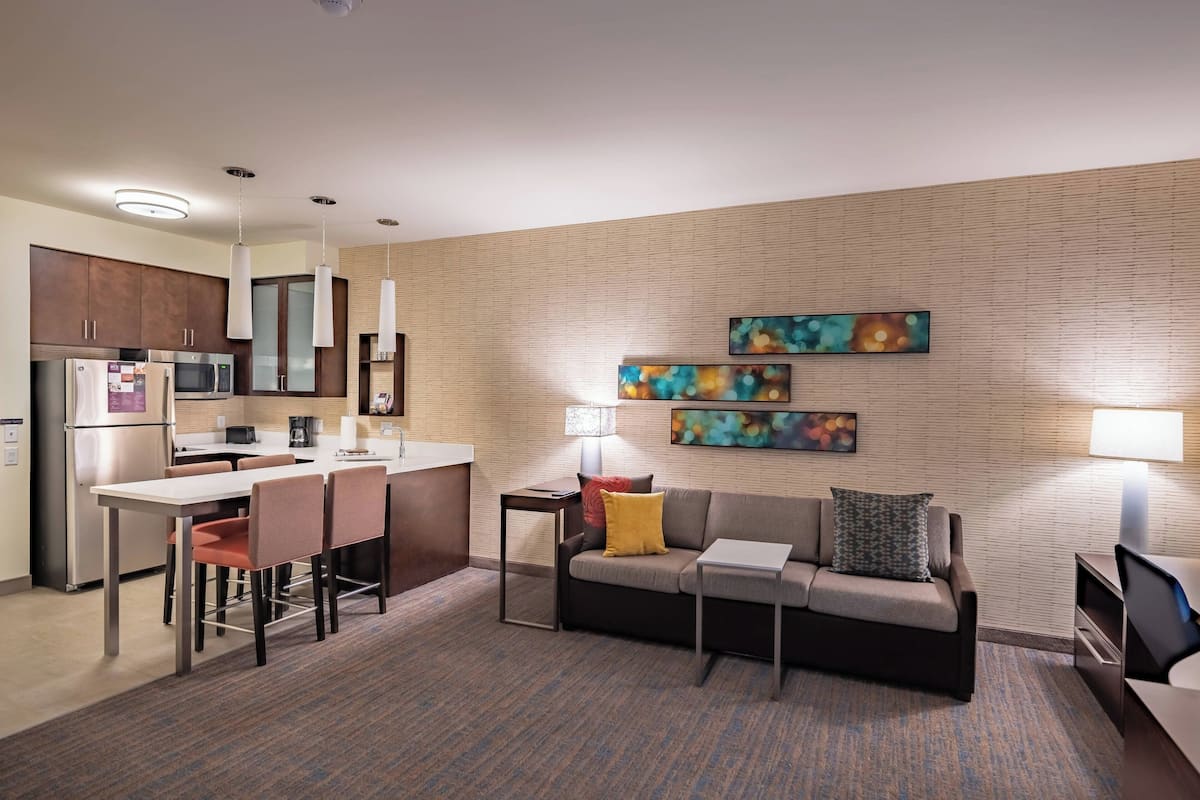 A modern hotel room with a kitchenette, a sofa, a coffee table, and artwork on the walls.