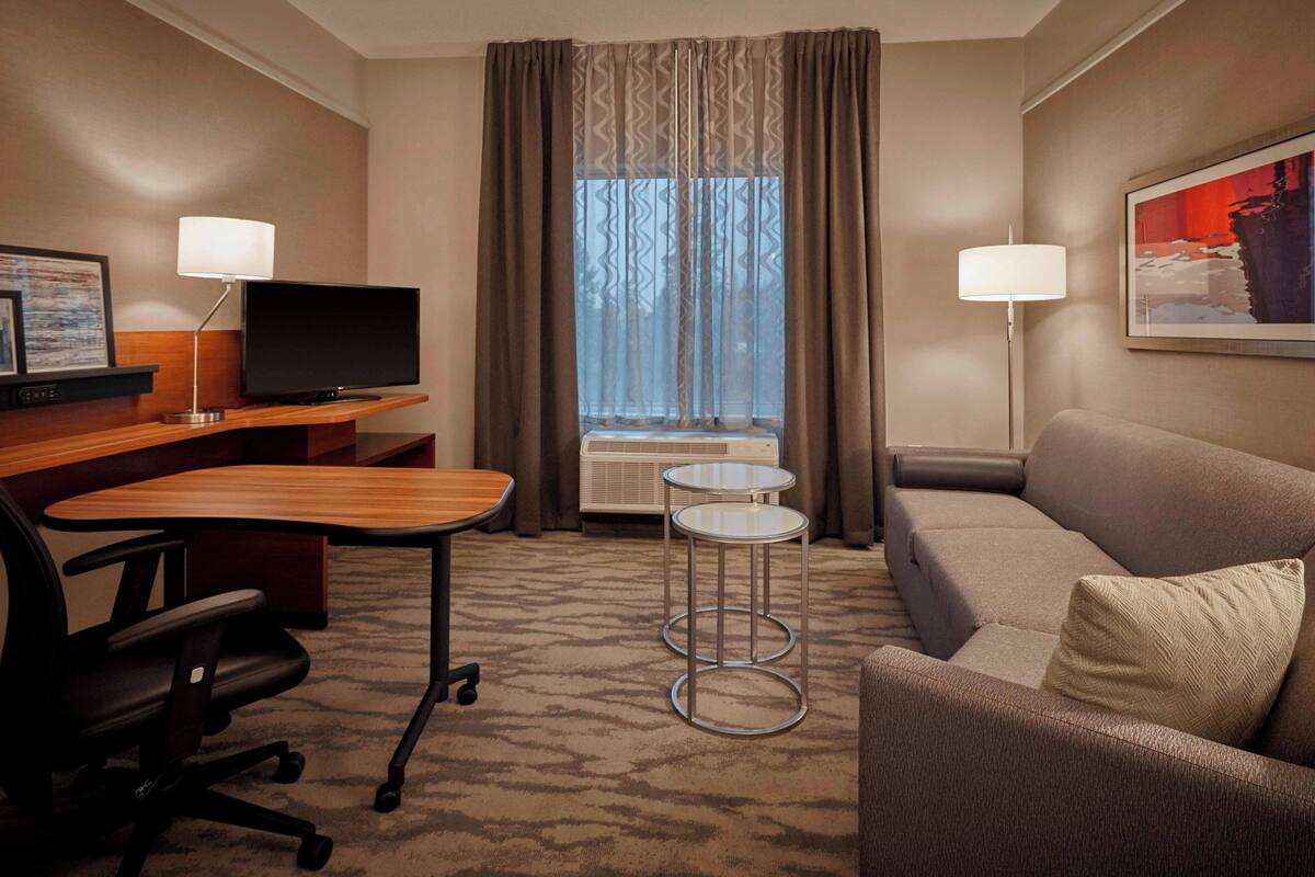Habitación de hotel con escritorio de madera, televisor, sofá, una mesita y una ventana con cortinas.