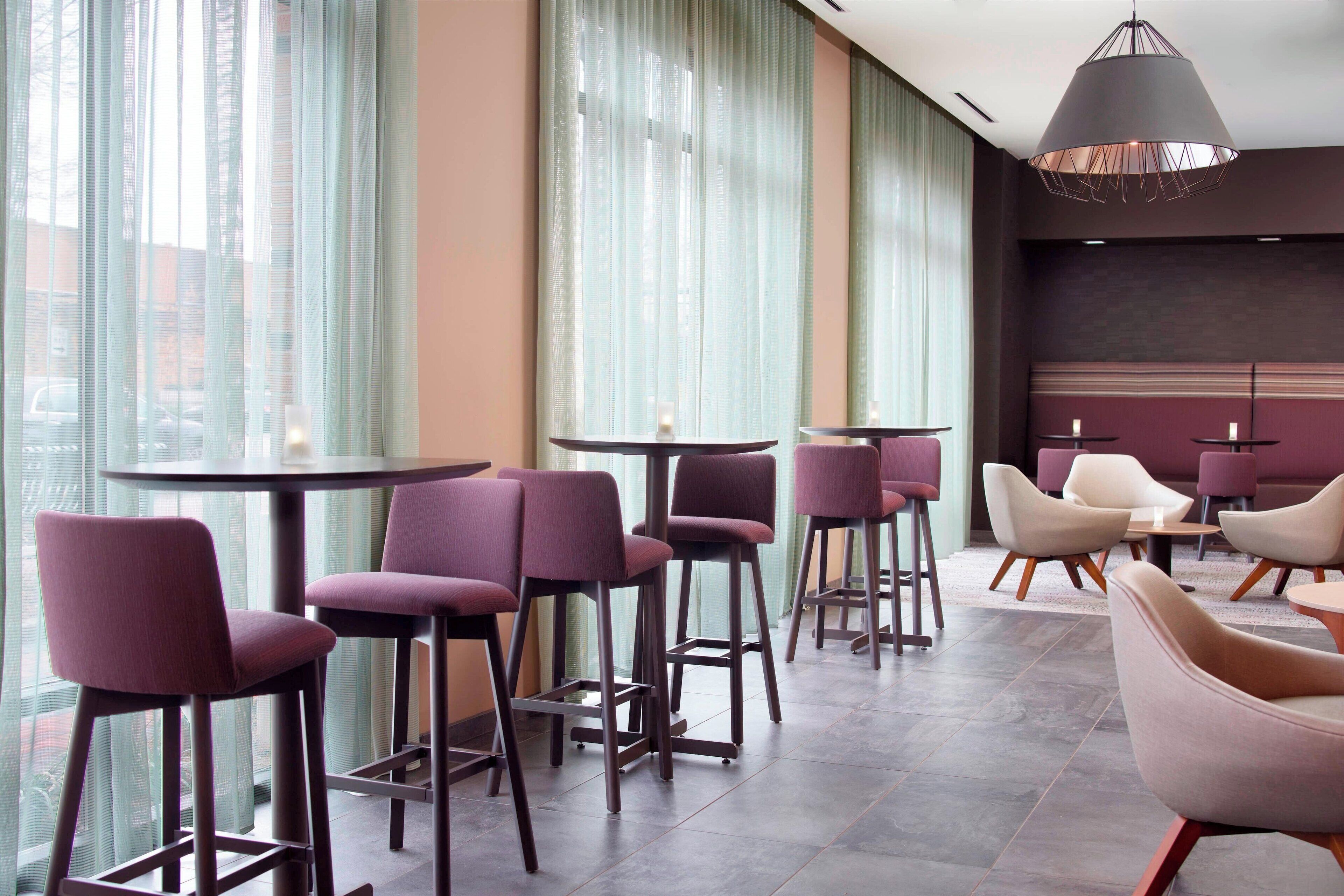 A modern bar area with high stools and round tables, large windows with sheer curtains, and a stylish pendant light.